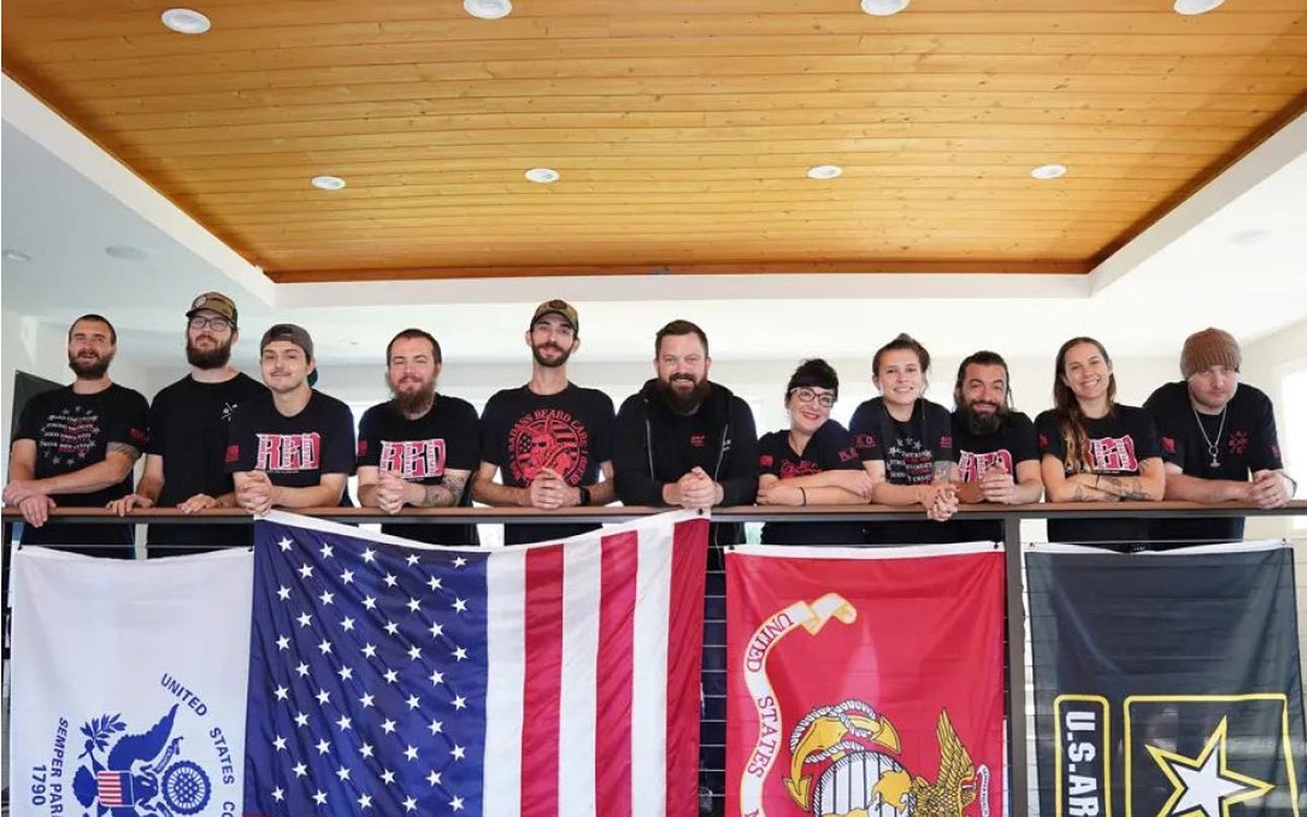 Ten Badass Beard Care team members standing together indoors, arms resting on a railing above four U.S. military-branch flags.